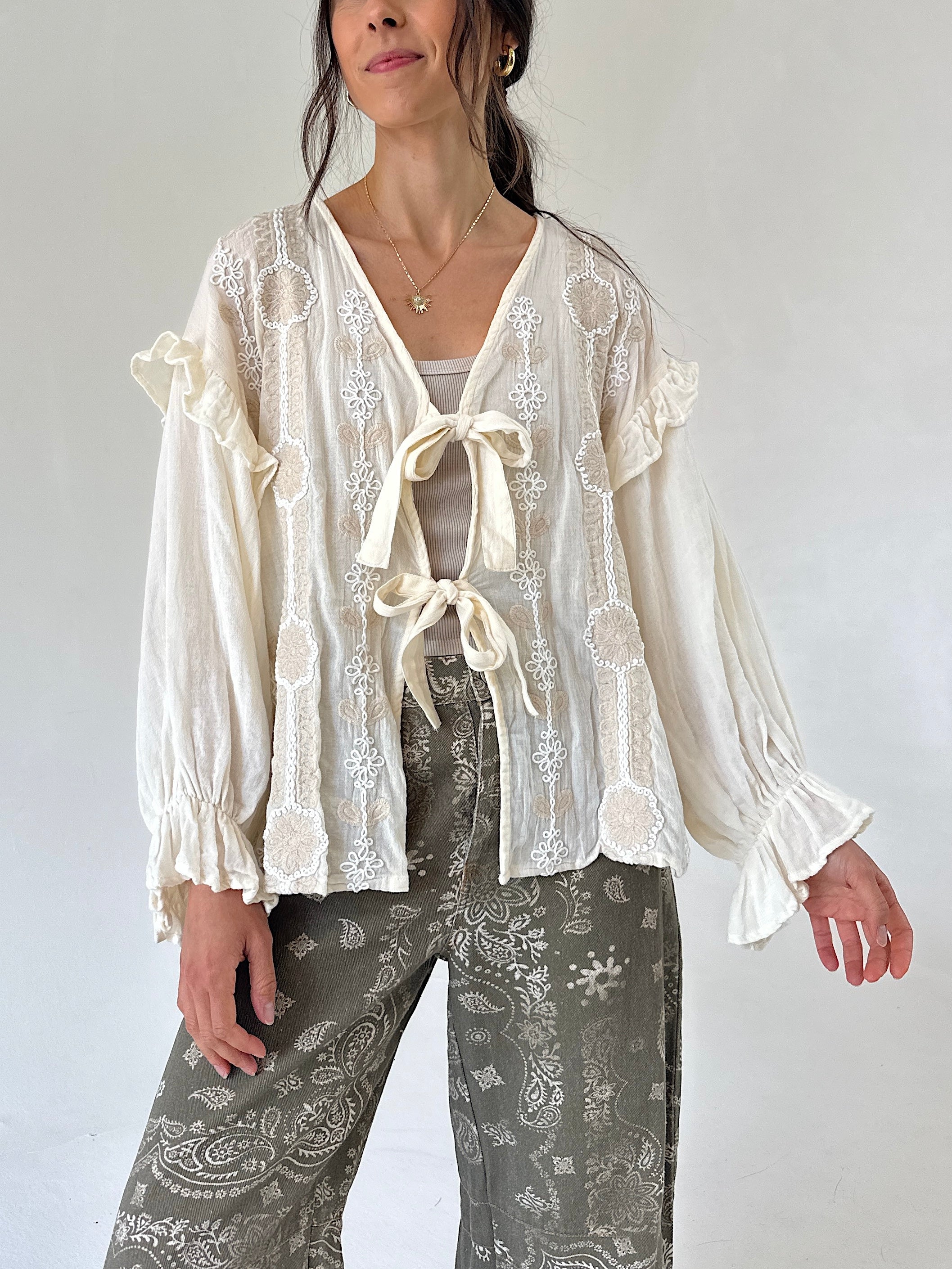 A woman wearing a cream-colored long-sleeve blouse with ruffled cuffs, lace and a front tie, paired with gray paisley pants, against a plain light background.