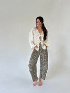 A woman modeling a bohemian-style outfit consisting of a cream lace blouse with ruffles and tie details, paired with olive green paisley bandana-print pants, standing against a plain light gray studio background.