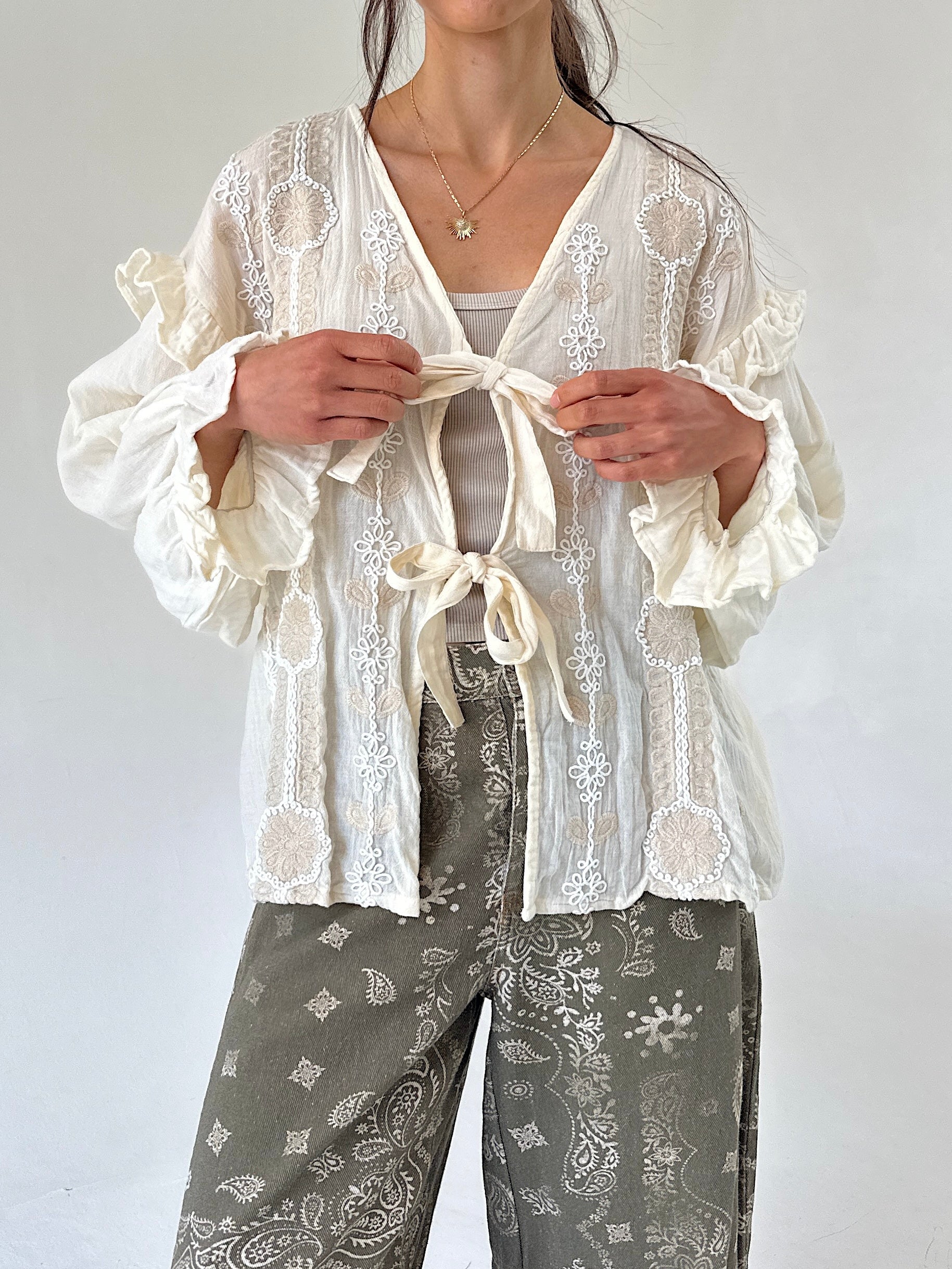 A woman wearing a cream tie-front long sleeve top with ruffled sleeves over a light gray top and gray paisley pants, photographed against a plain light background.