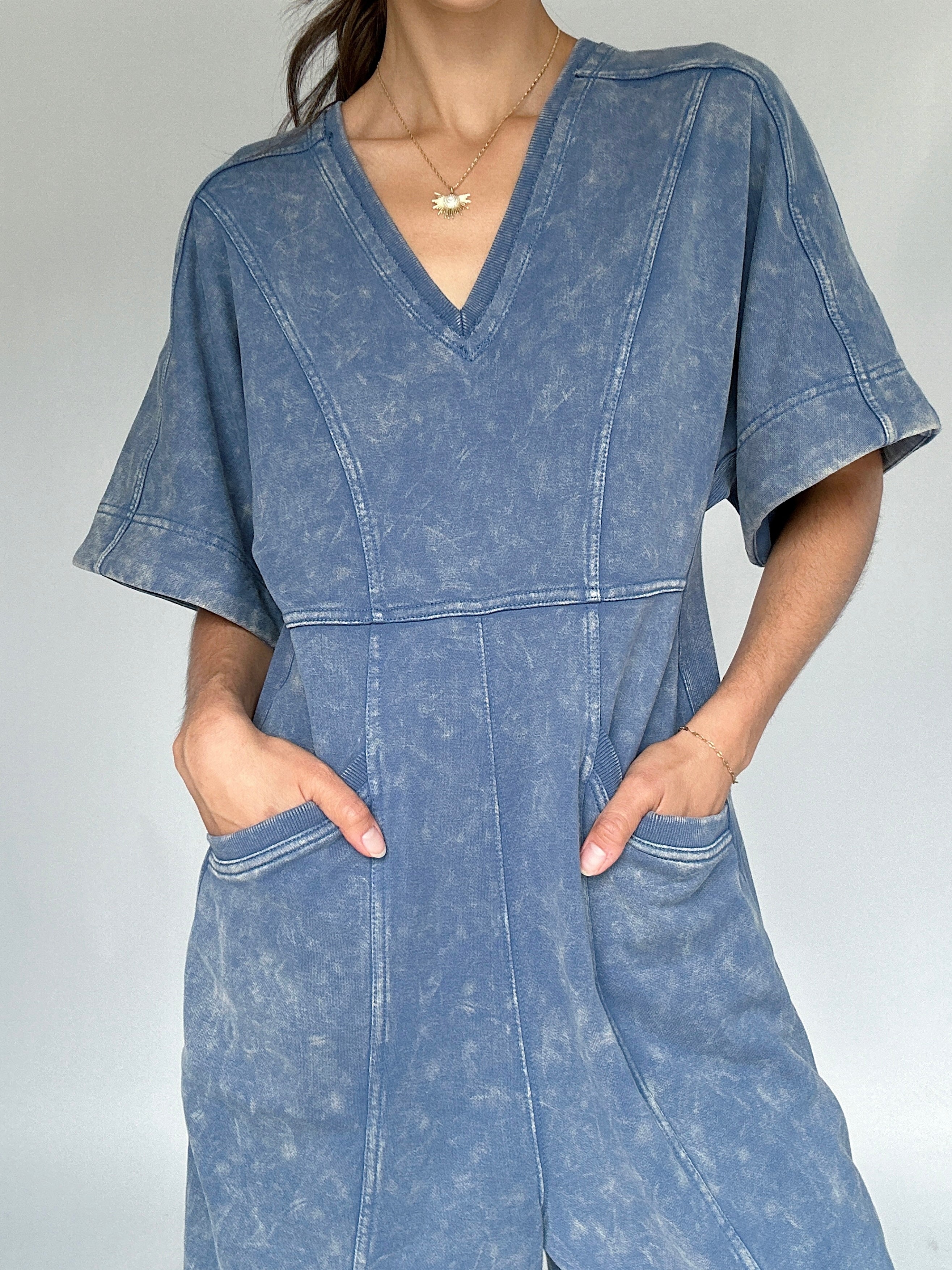 Person wearing a blue mineral-wash jumpsuit against a plain background up close to show v-neckline and pockets