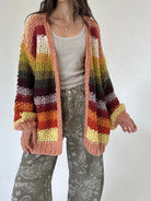 A person wearing a chunky knit open-front cardigan with horizontal multicolor stripes in peach, rust, orange, yellow, olive, and dark brown, layered over a light gray top and gray paisley pants against a plain light background.