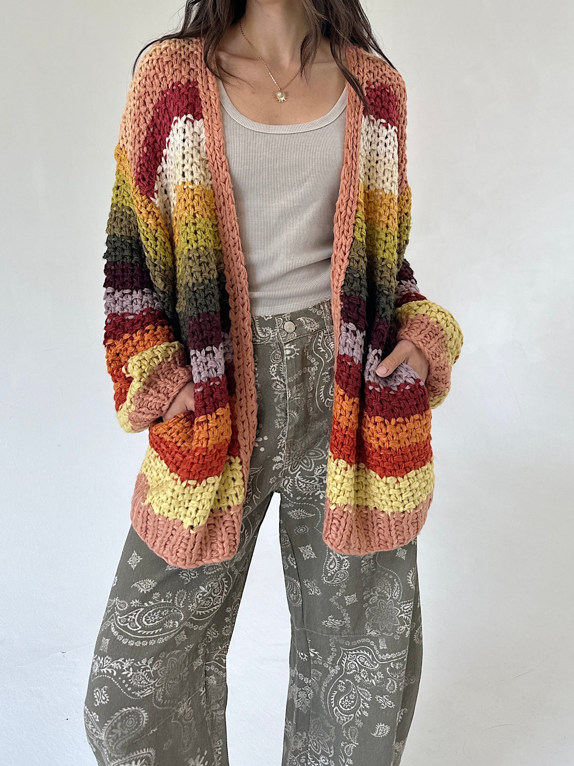 A person wearing a chunky striped crochet cardigan with horizontal bands of cream, yellow, orange, red, and brown, layered over a light gray tank top and gray paisley pants, against a plain off-white background.