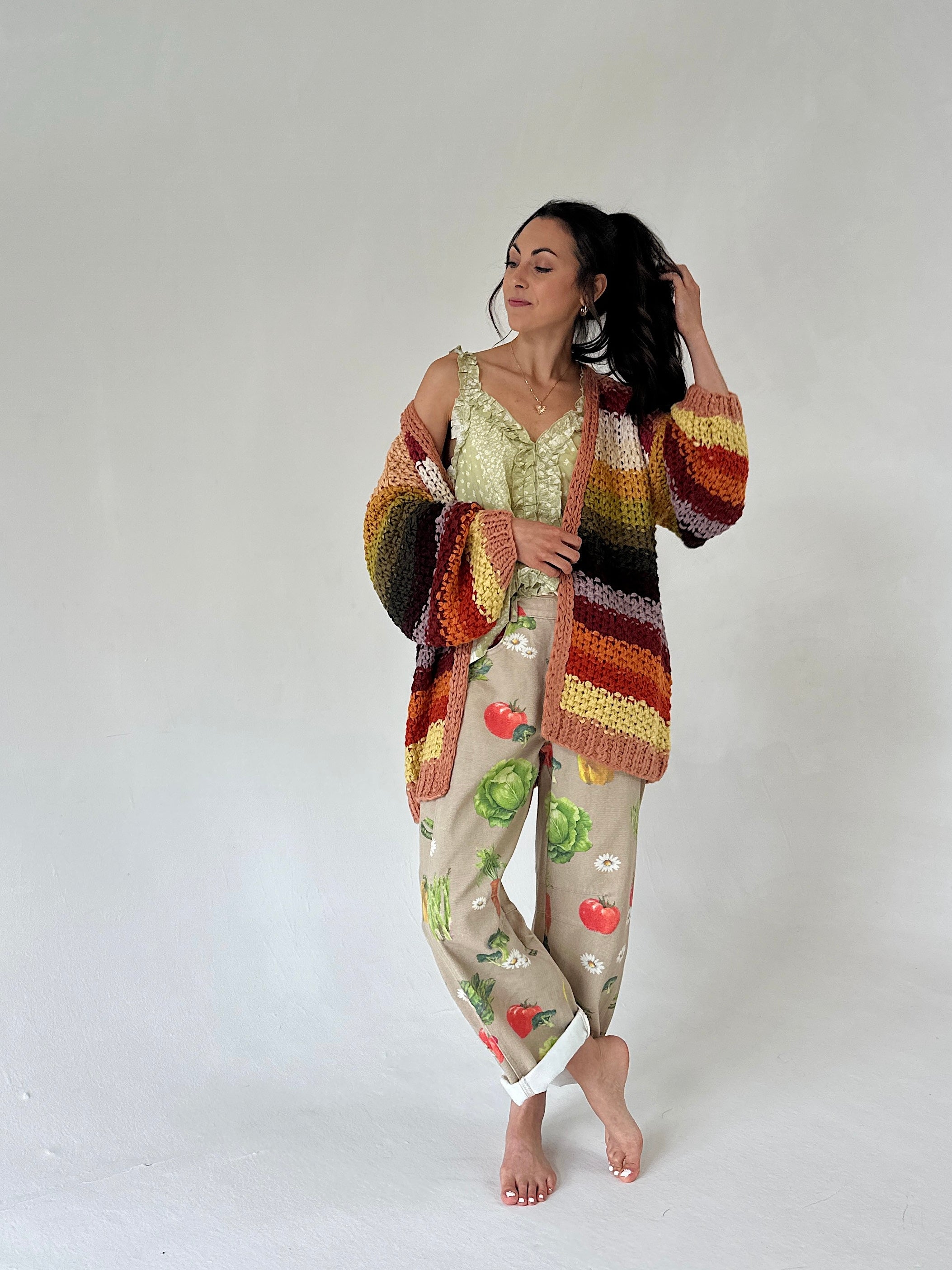 A woman stands barefoot against a plain light gray studio background wearing barrel pants with a vegetable print, a striped crochet cardigan in earthy tones, and a pale green lace-trim camisole.