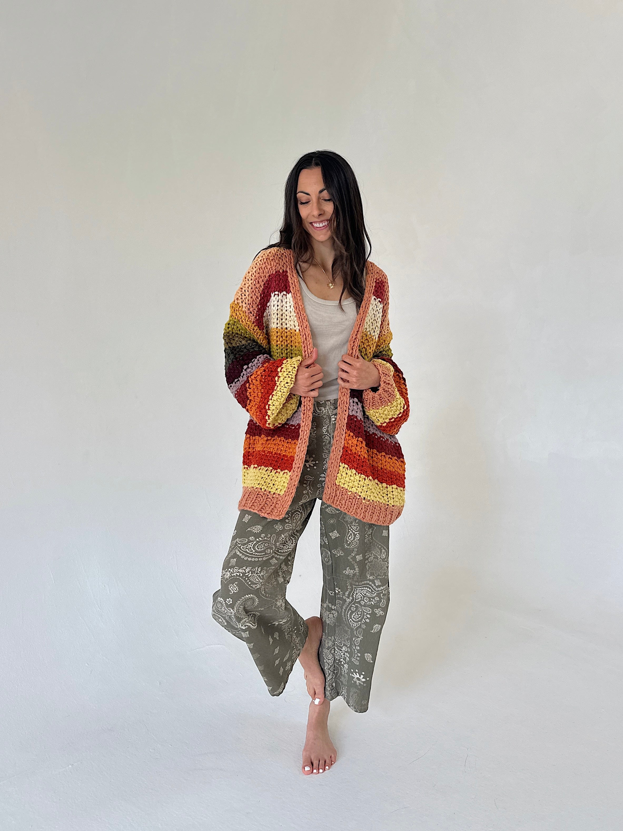 A woman stands barefoot against a plain light background wearing a chunky multicolor striped cardigan with rust, orange, yellow, and brown tones, a light beige top, and loose sage green bandana patterned barrel pants.