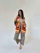 A woman stands barefoot against a plain light background wearing a chunky multicolor striped cardigan with rust, orange, yellow, and brown tones, a light beige top, and loose sage green bandana patterned barrel pants.