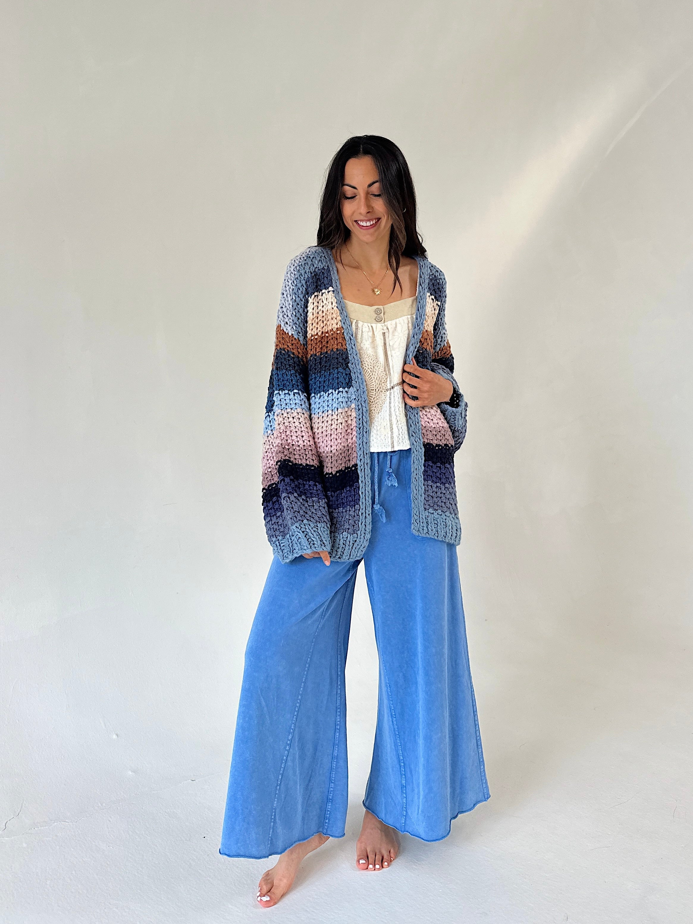 A woman stands barefoot against a plain light background wearing a chunky multicolor striped knit cardigan over a white top and wide-leg blue pants.