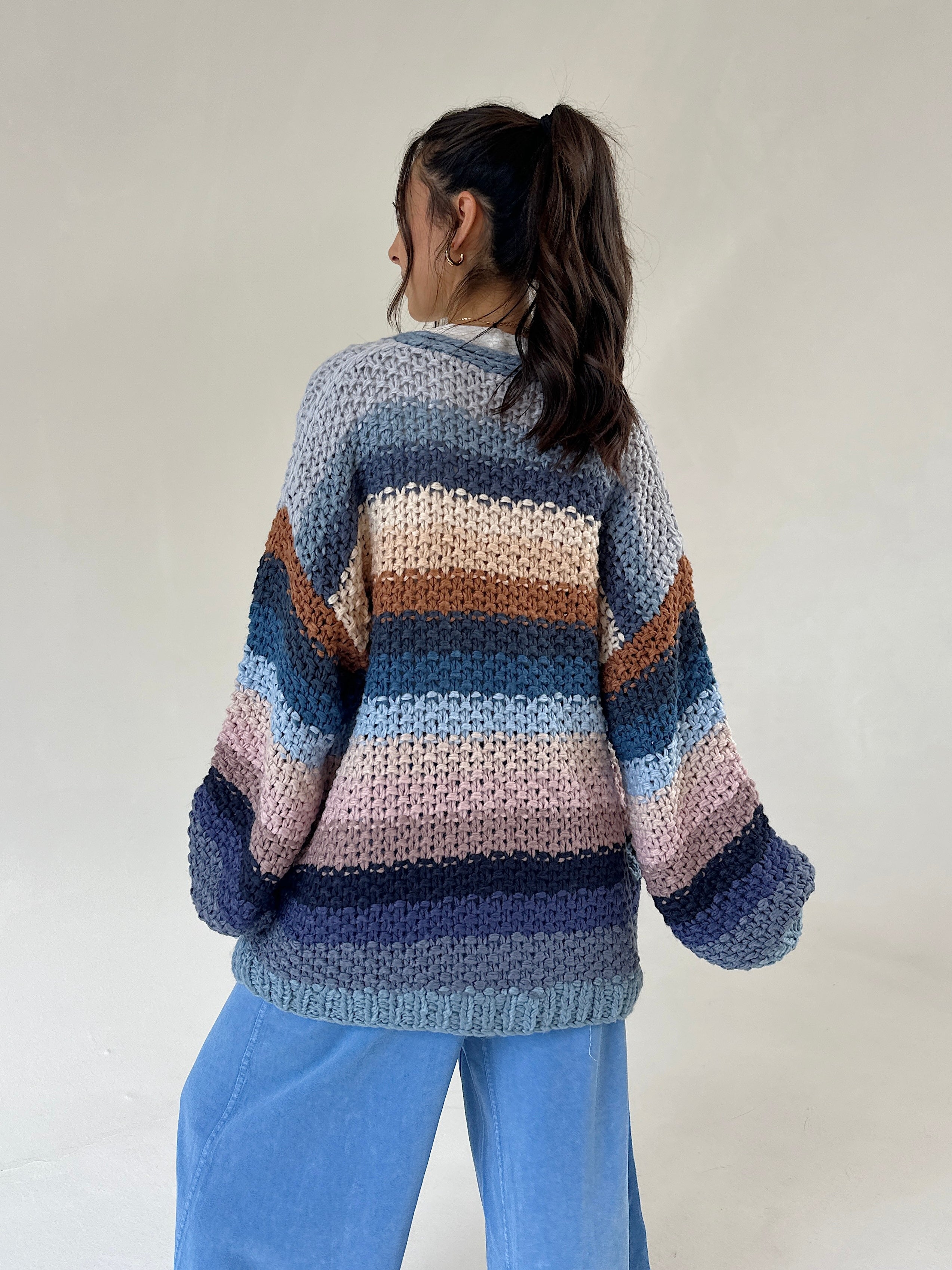 A person shown from the back wearing a chunky knit striped sweater with horizontal bands of blue, navy, beige, pink, and brown, paired with light blue pants, against a plain off-white background.