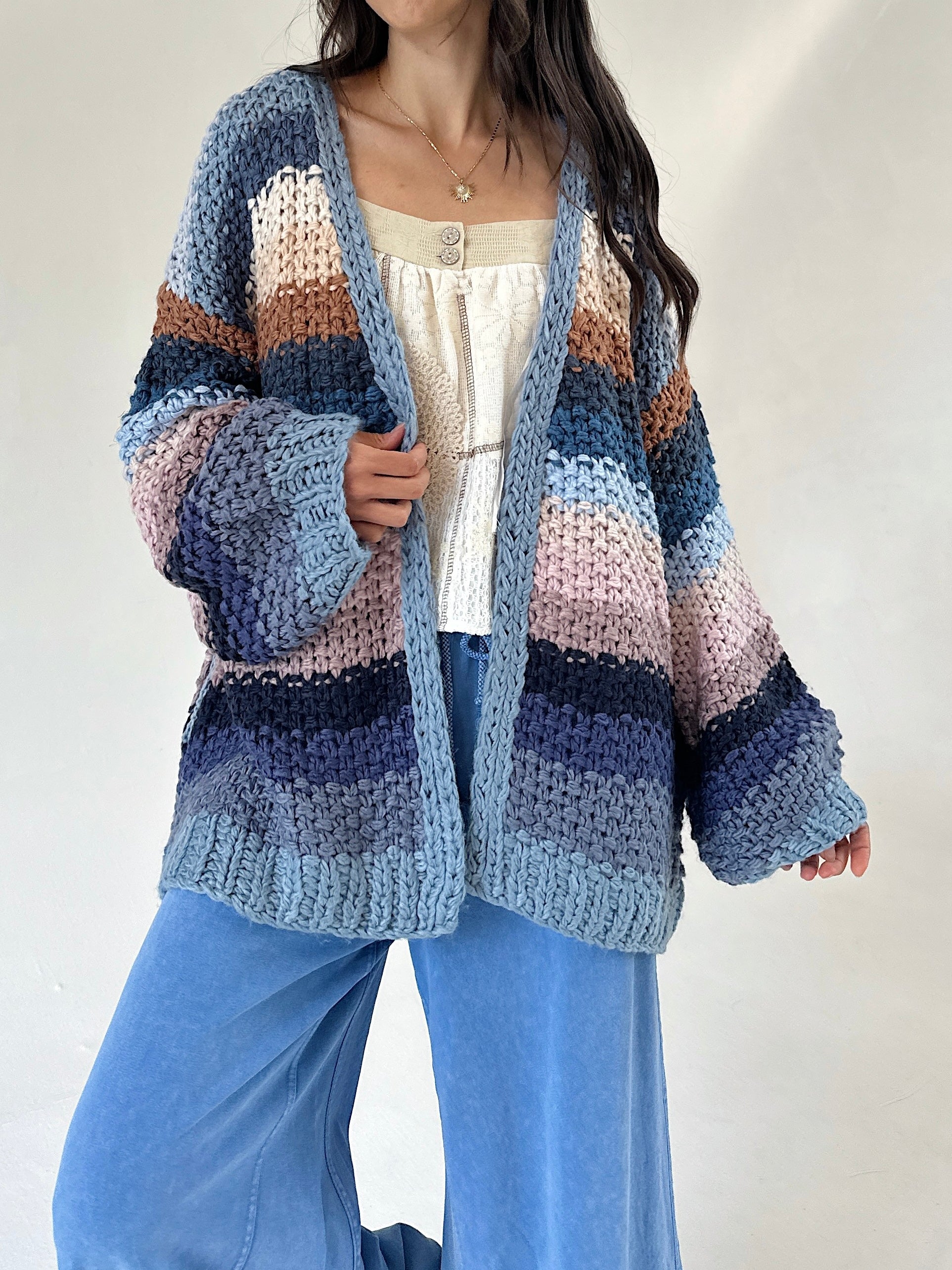 A woman wearing a chunky knit open-front cardigan with horizontal stripes in shades of blue, navy, beige, pink, and brown, styled over a cream top and blue wide-leg pants.
