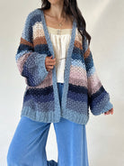 A woman wearing a chunky knit open-front cardigan with horizontal stripes in shades of blue, navy, beige, pink, and brown, styled over a cream top and blue wide-leg pants.