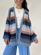A woman wearing a chunky knit open-front cardigan with horizontal stripes in shades of blue, navy, pink, beige, and brown over a white top and blue pants, photographed against a plain light background.