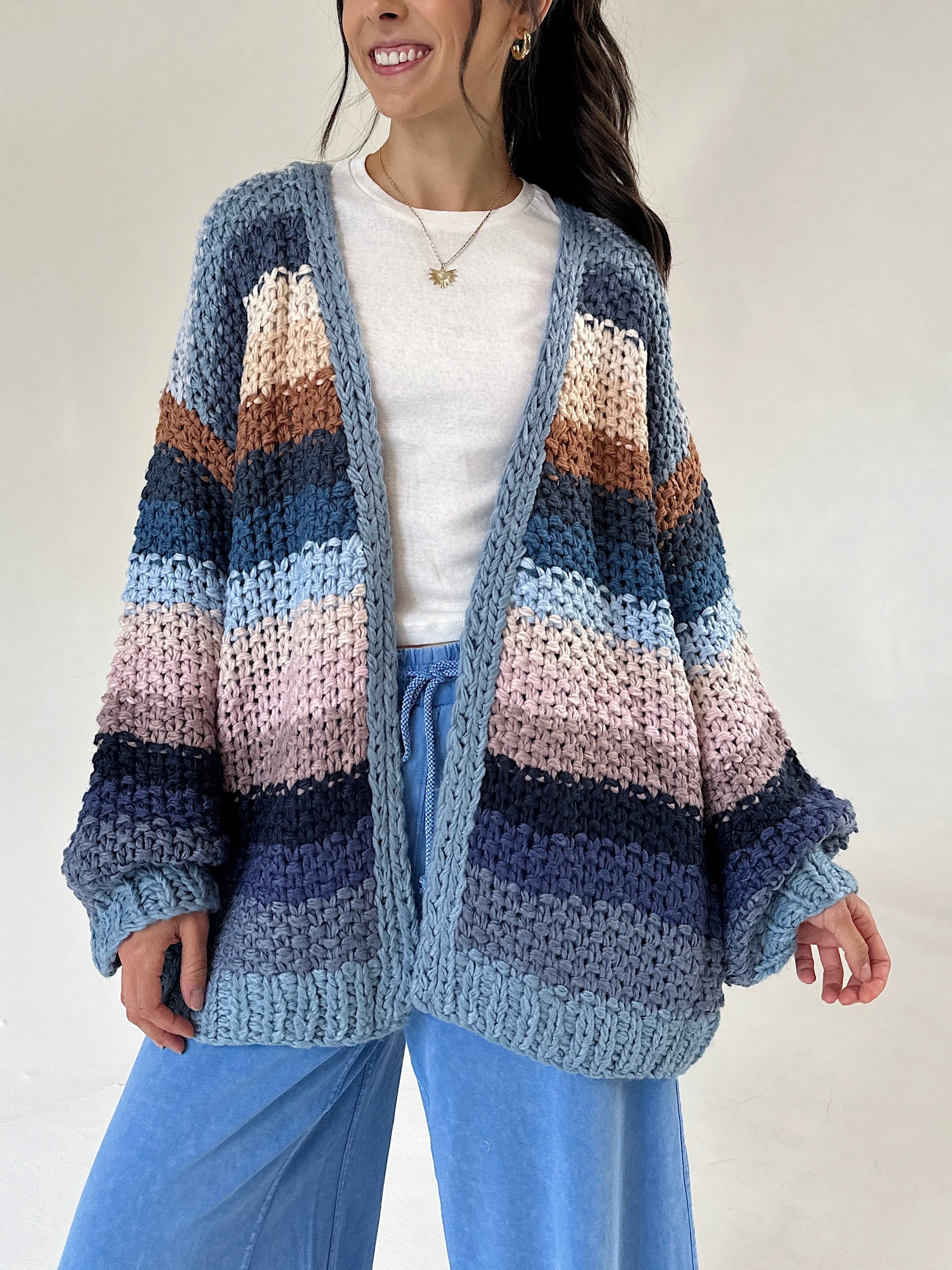A woman wearing a chunky knit open-front cardigan with horizontal stripes in shades of blue, navy, pink, beige, and brown over a white top and blue pants, photographed against a plain light background.