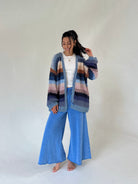 A woman stands barefoot against a plain light gray studio background, wearing a chunky knit cardigan with horizontal stripes in blue, purple, pink, and brown over a white top and wide-leg blue pants.