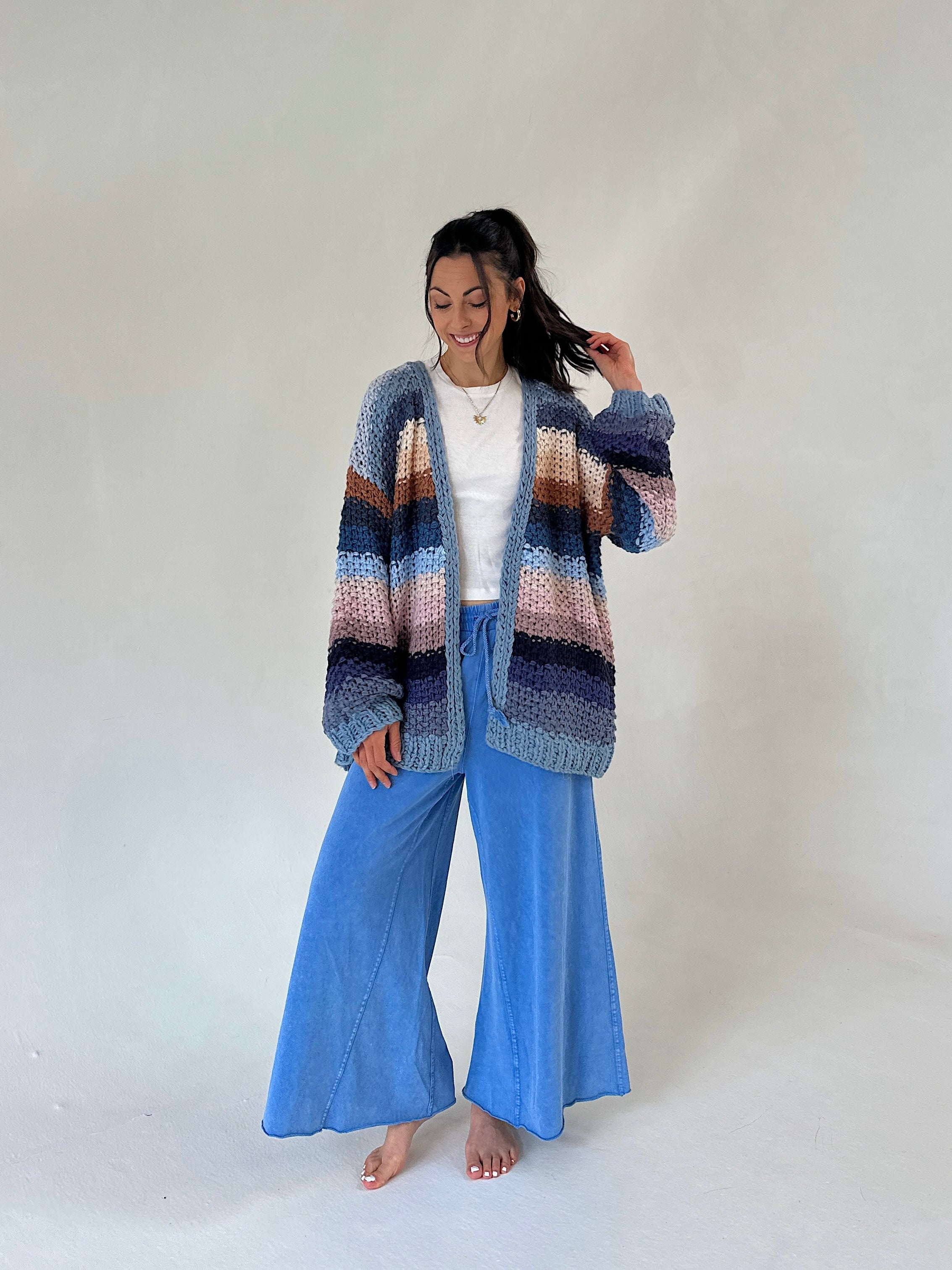 A woman stands barefoot against a plain light gray studio background, wearing a chunky knit cardigan with horizontal stripes in blue, purple, pink, and brown over a white top and wide-leg blue pants.