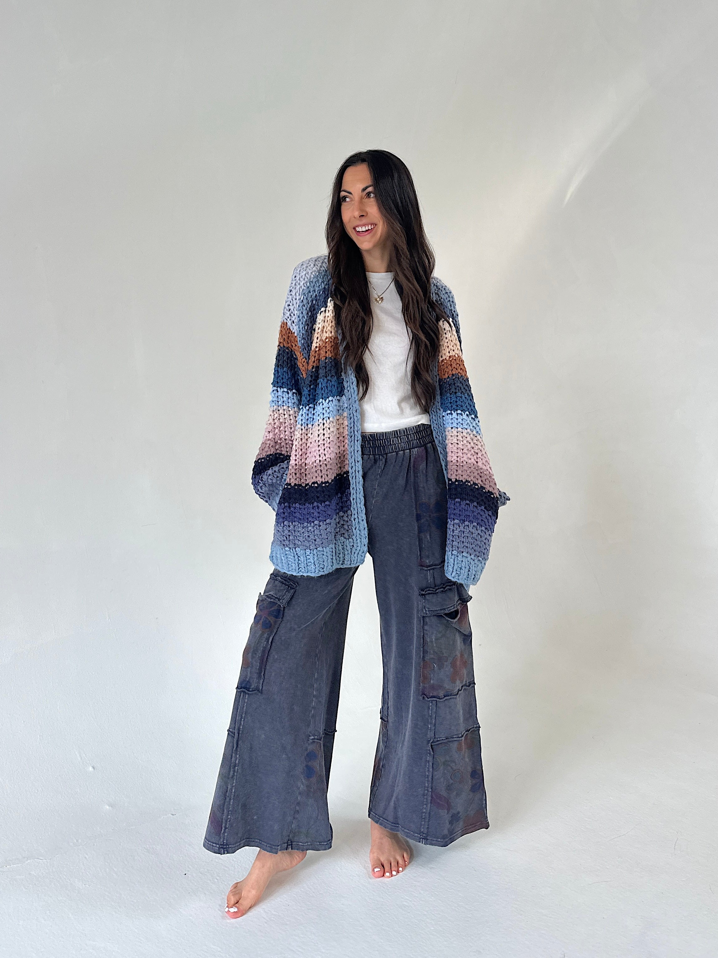 A woman stands barefoot against a plain light gray studio backdrop, wearing a loose multicolor striped knit cardigan over a white top and dark gray wide-leg cargo pants.