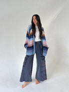 A woman stands barefoot against a plain light gray studio backdrop, wearing a loose multicolor striped knit cardigan over a white top and dark gray wide-leg cargo pants.