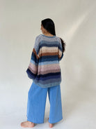 A woman stands barefoot against a plain light background, viewed from the back, wearing a loose striped knit sweater with blue wide-leg pants.