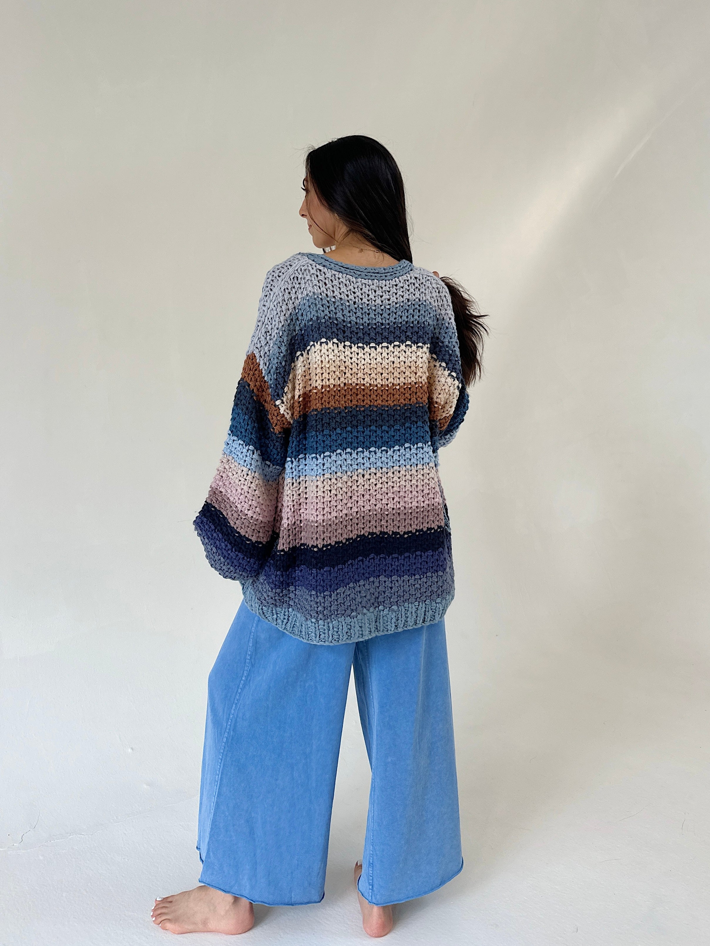 A woman stands barefoot against a plain light background, viewed from the back, wearing a loose striped knit sweater with blue wide-leg pants.