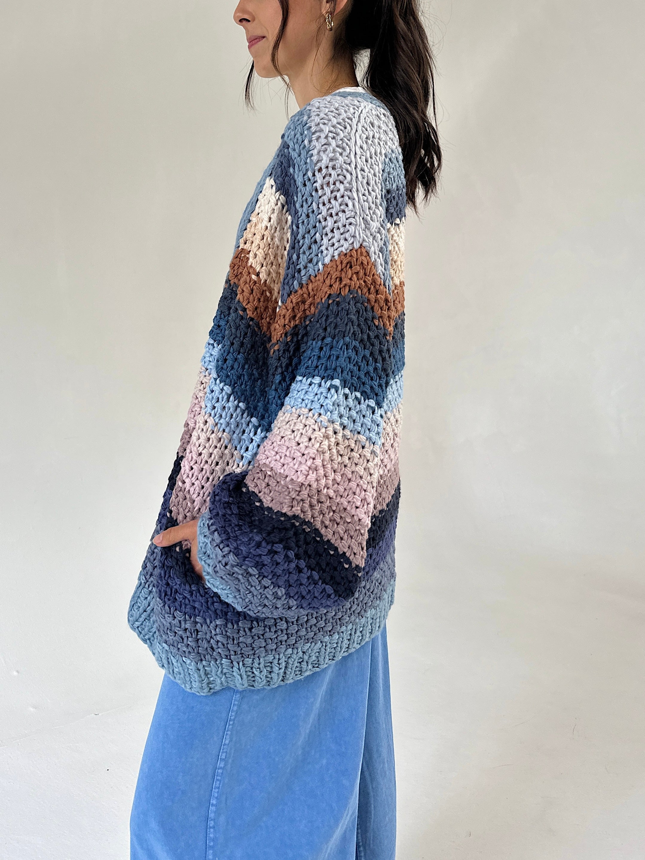 A woman wearing a chunky cardigan with horizontal stripes in shades of blue, navy, pink, and brown, paired with light blue jeans against a plain off-white background.