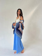 A woman stands barefoot against a plain light gray studio background, wearing blue wide leg pants and holding a chunky knit cardigan draped over her shoulders. The cardigan has a striped pattern in shades of blue, navy, beige, and pink.