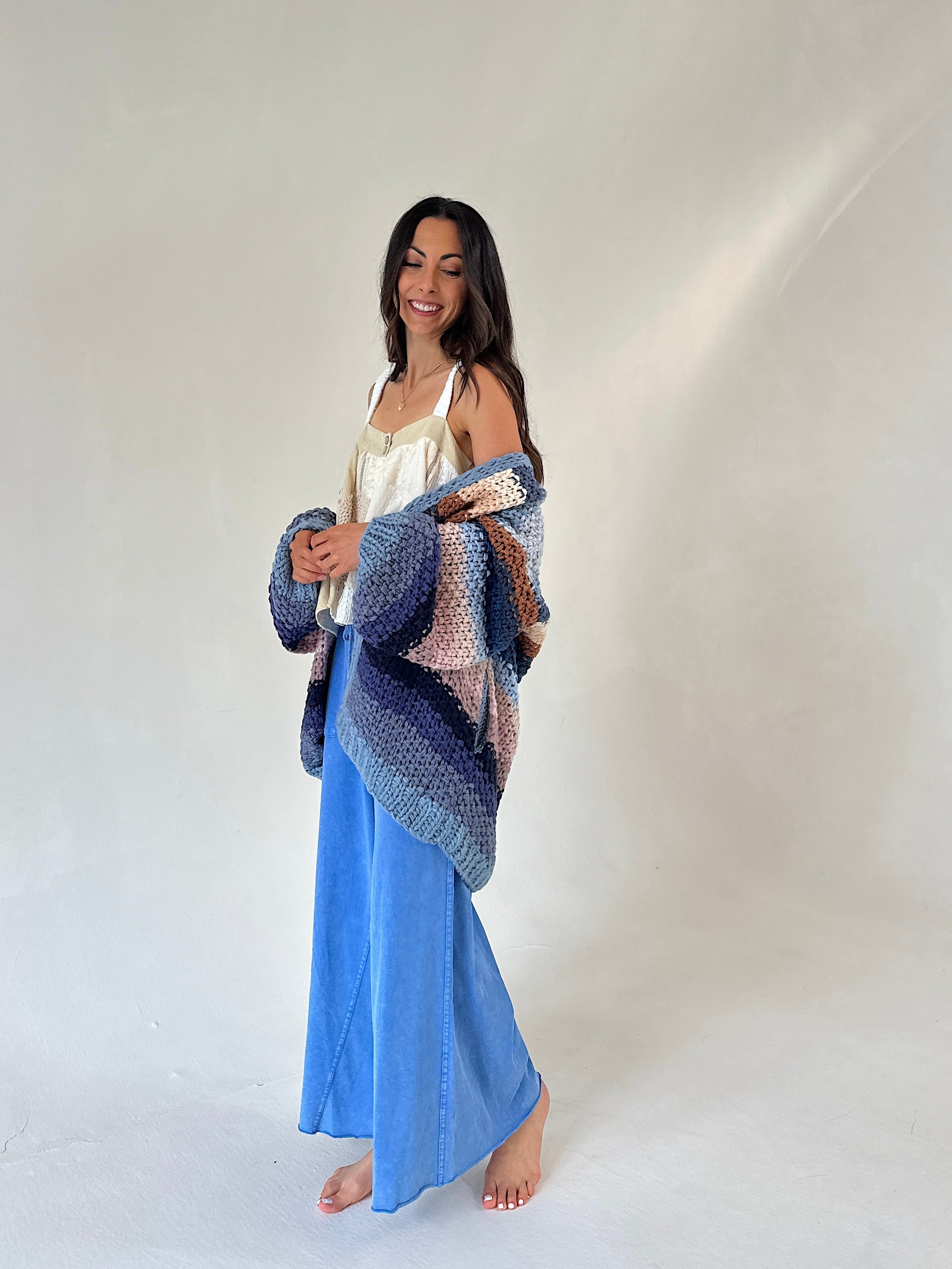 A woman stands barefoot against a plain light gray studio background, wearing blue wide leg pants and holding a chunky knit cardigan draped over her shoulders. The cardigan has a striped pattern in shades of blue, navy, beige, and pink.