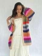 Woman wearing a colorful striped crochet cardigan over a white dress against a plain background