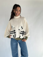 Woman wearing a cream turtle neck sweater with sheep jumping over a fence pattern on a plain background