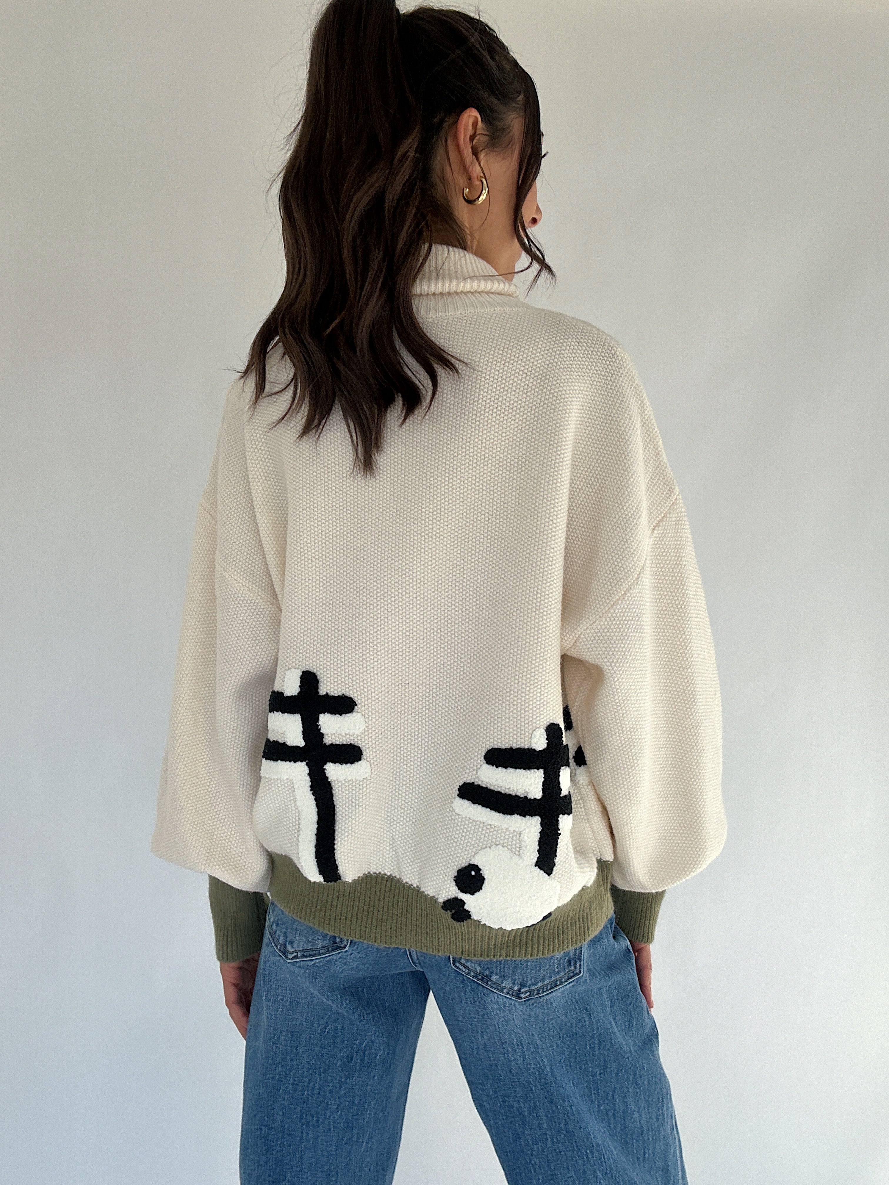 Person wearing a cream sweater with sheep and a fence on the back, standing against a white background