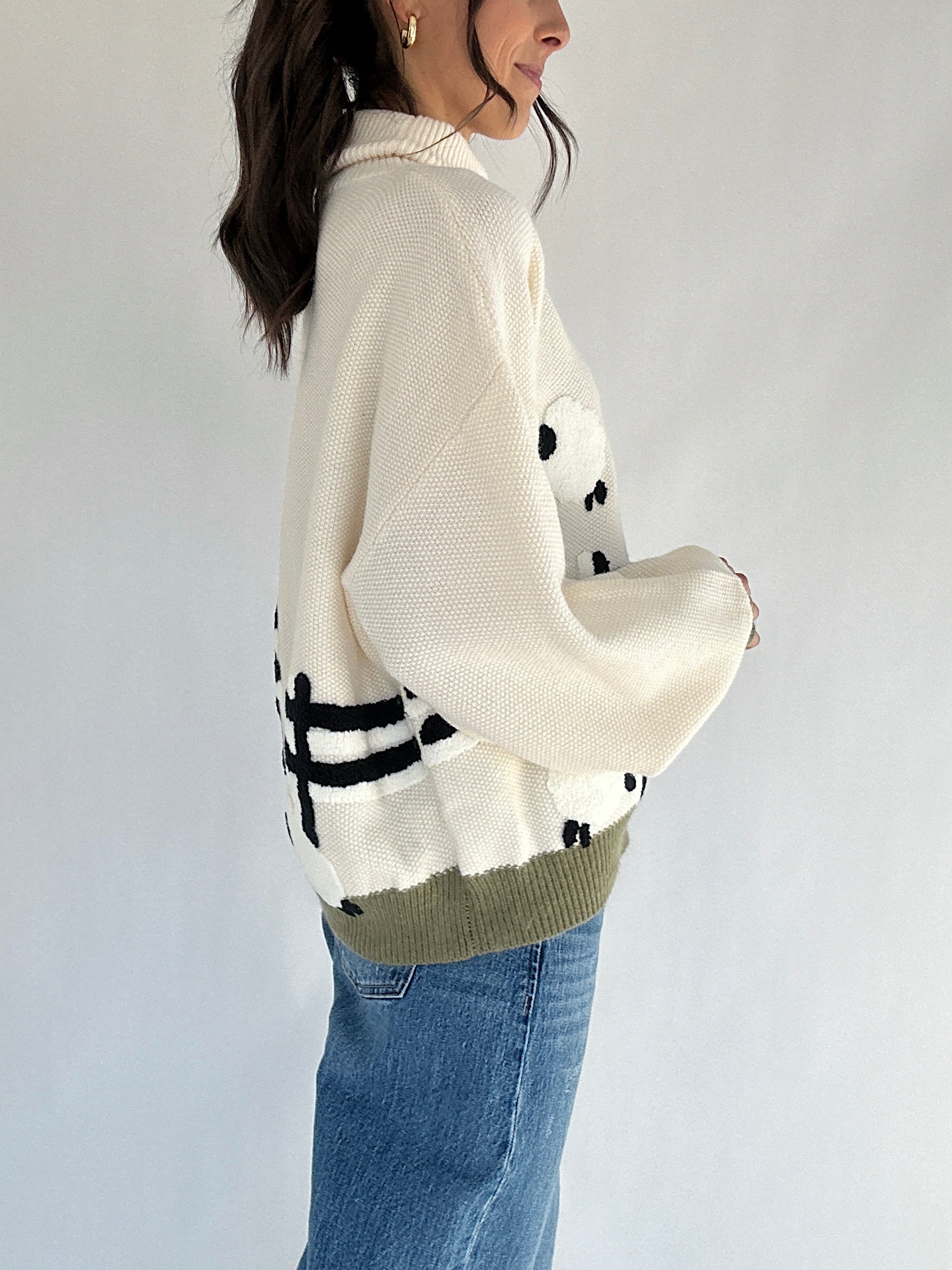 Person wearing a cream turtle neck sweater with graphic patterns and blue jeans on a plain background showing side