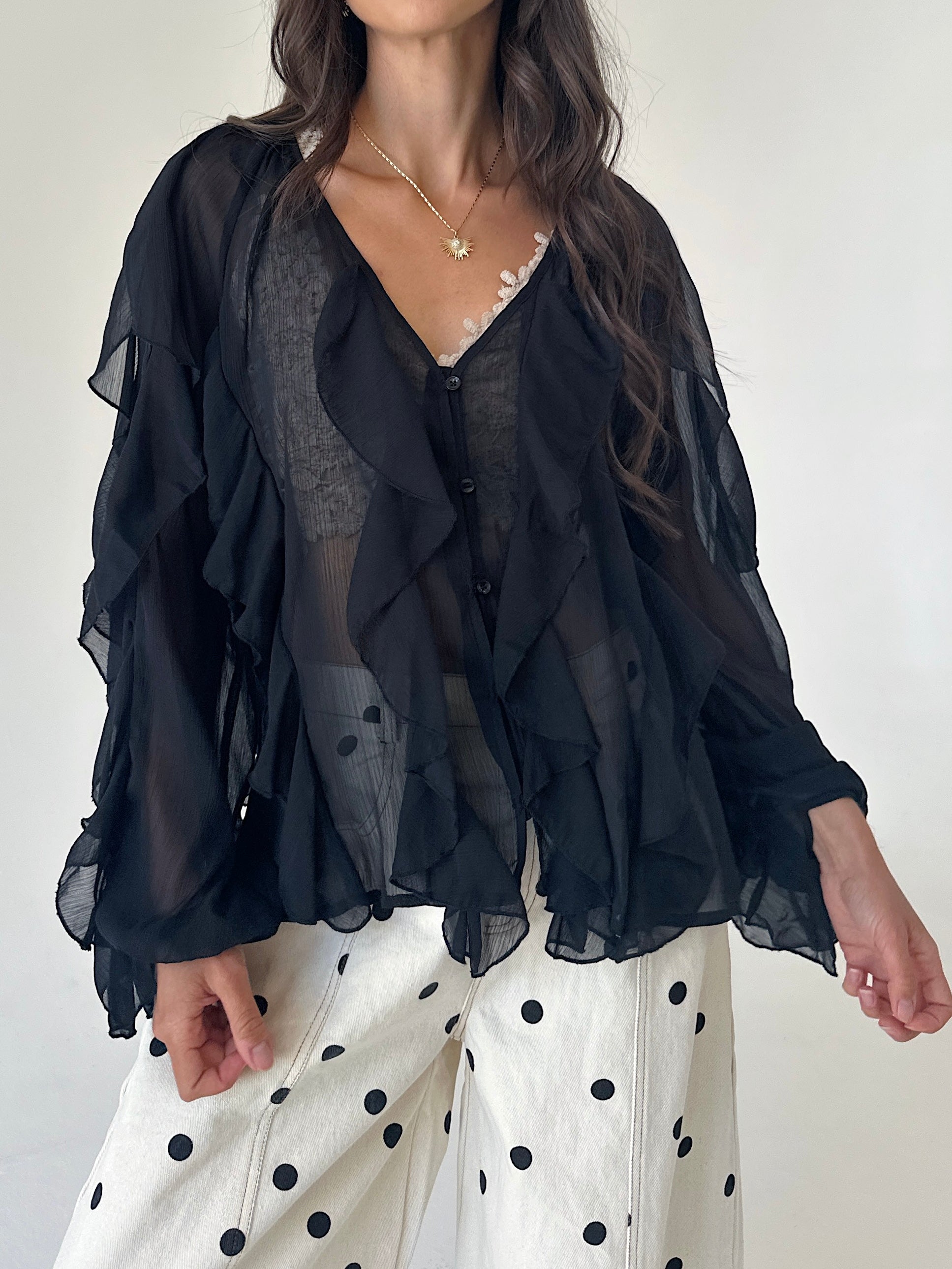 A woman wearing a sheer black ruffled blouse over a gray lace top, paired with white pants featuring black polka dots, against a plain off-white background.