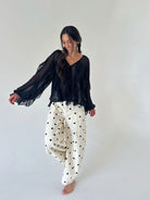A woman modeling a black sheer long-sleeve blouse and cream wide-leg pants with black polka dots, standing against a plain light gray studio background.