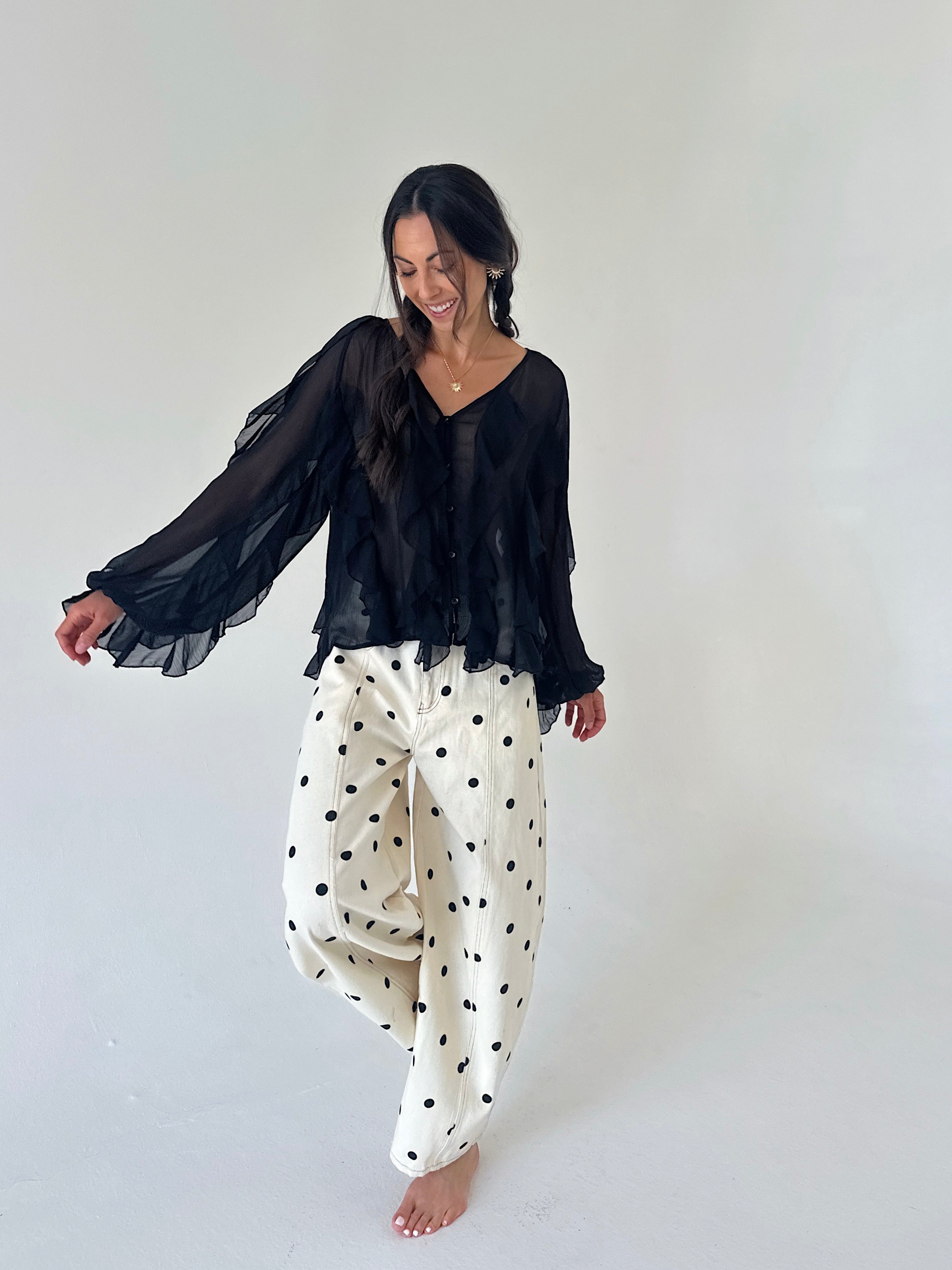 A woman modeling a black sheer long-sleeve blouse and cream wide-leg pants with black polka dots, standing against a plain light gray studio background.