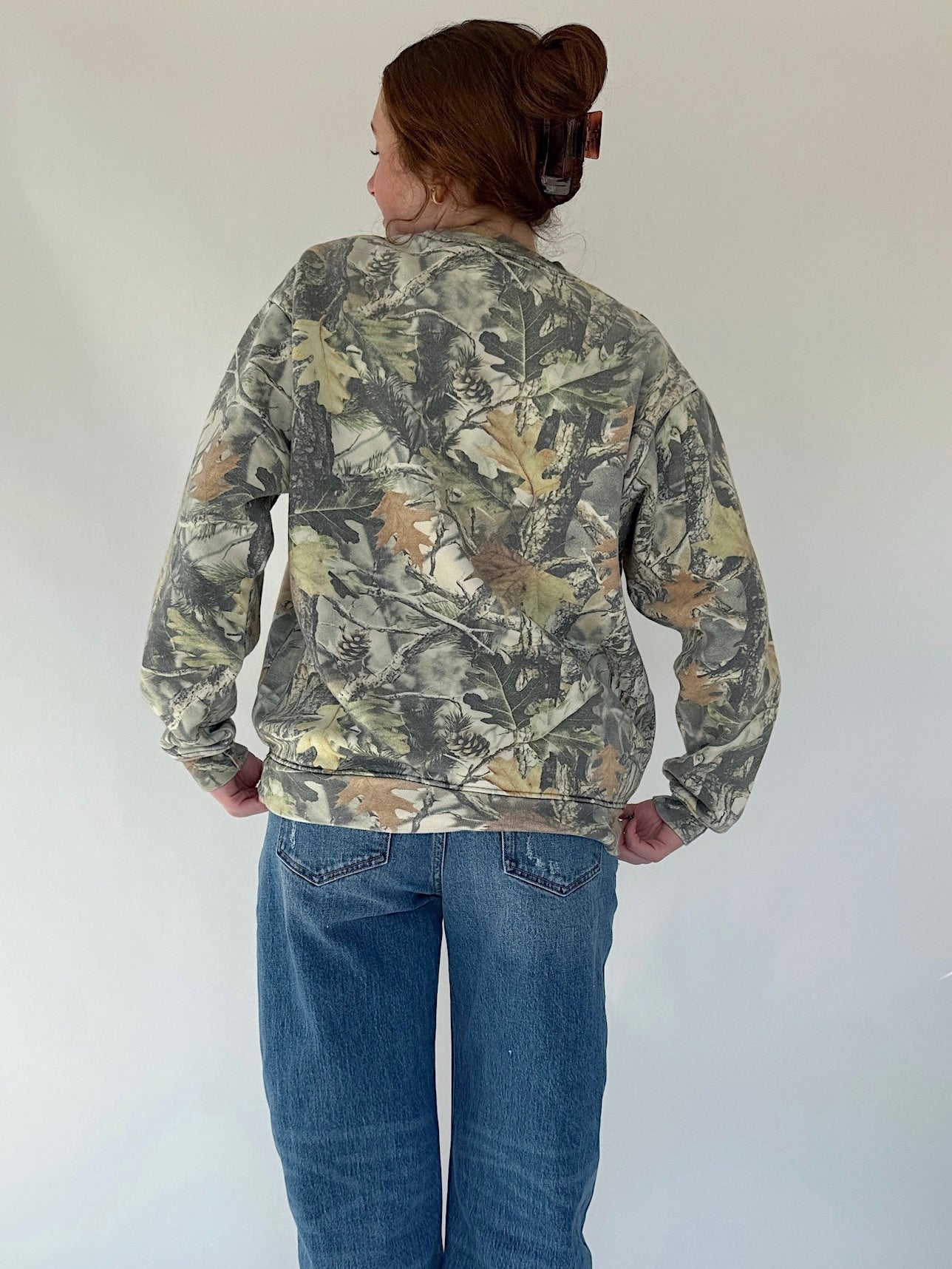 Person wearing a camouflage sweatshirt and blue jeans against a white background showing back