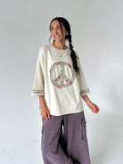 A woman modeling a cream-colored oversized sweatshirt with a colorful peace sign graphic on the front, paired with wide-leg mauve cargo pants, against a plain light gray background.