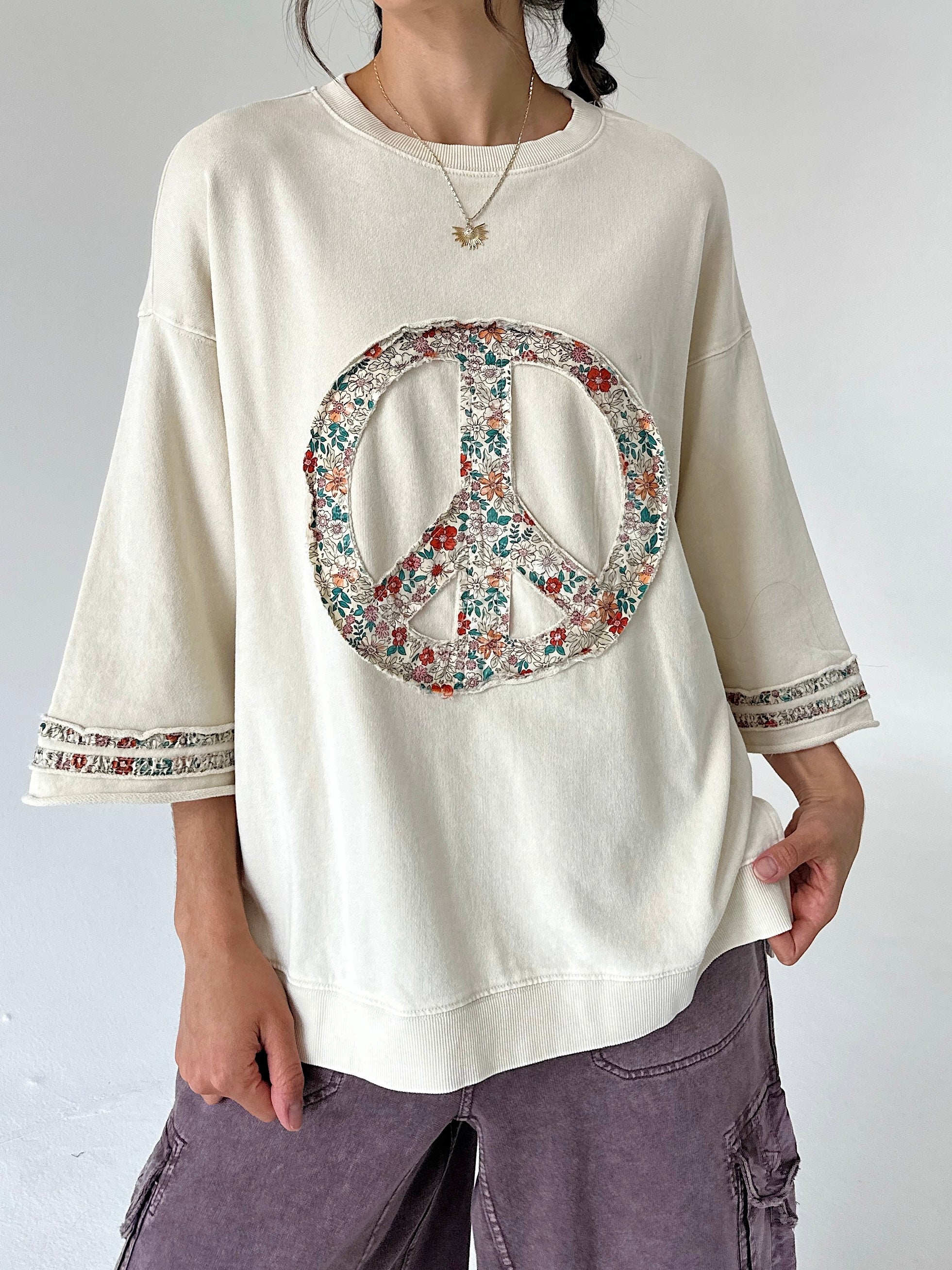 A person wearing an oversized cream sweatshirt with a large peace sign graphic on the front, filled with a floral pattern in red, green, and blue. The sweatshirt has 3/4 sleeves with patterned cuffs.