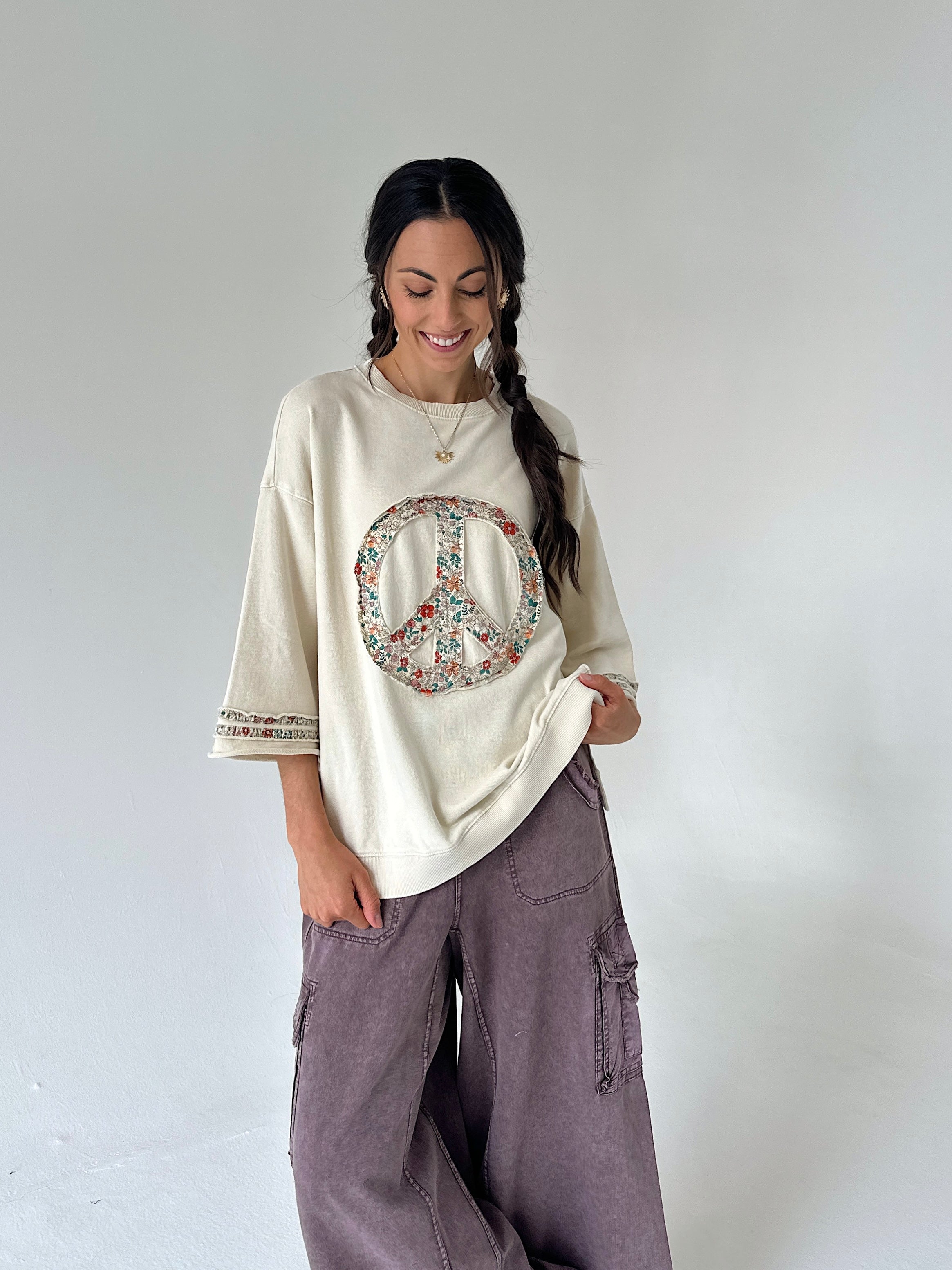 A woman modeling a cream-colored oversized t-shirt with a colorful peace sign graphic on the front, paired with wide-leg mauve cargo pants, against a plain light gray studio background.