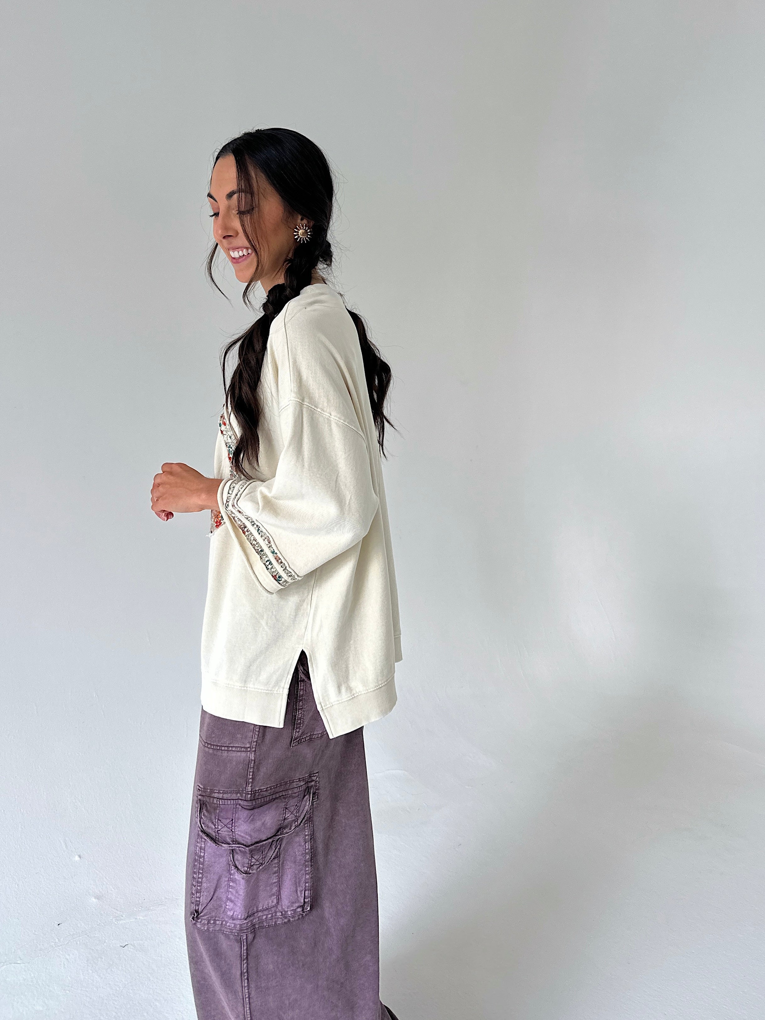 A woman wearing a cream-colored knit sweater with embroidered details on the sleeve and a purple cargo skirt, standing against a plain light gray studio background.