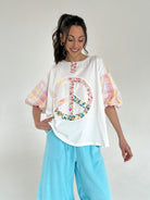 Woman wearing a white tee with a peace symbol design and colorful plaid short sleeves, paired with light blue pants on a plain background.