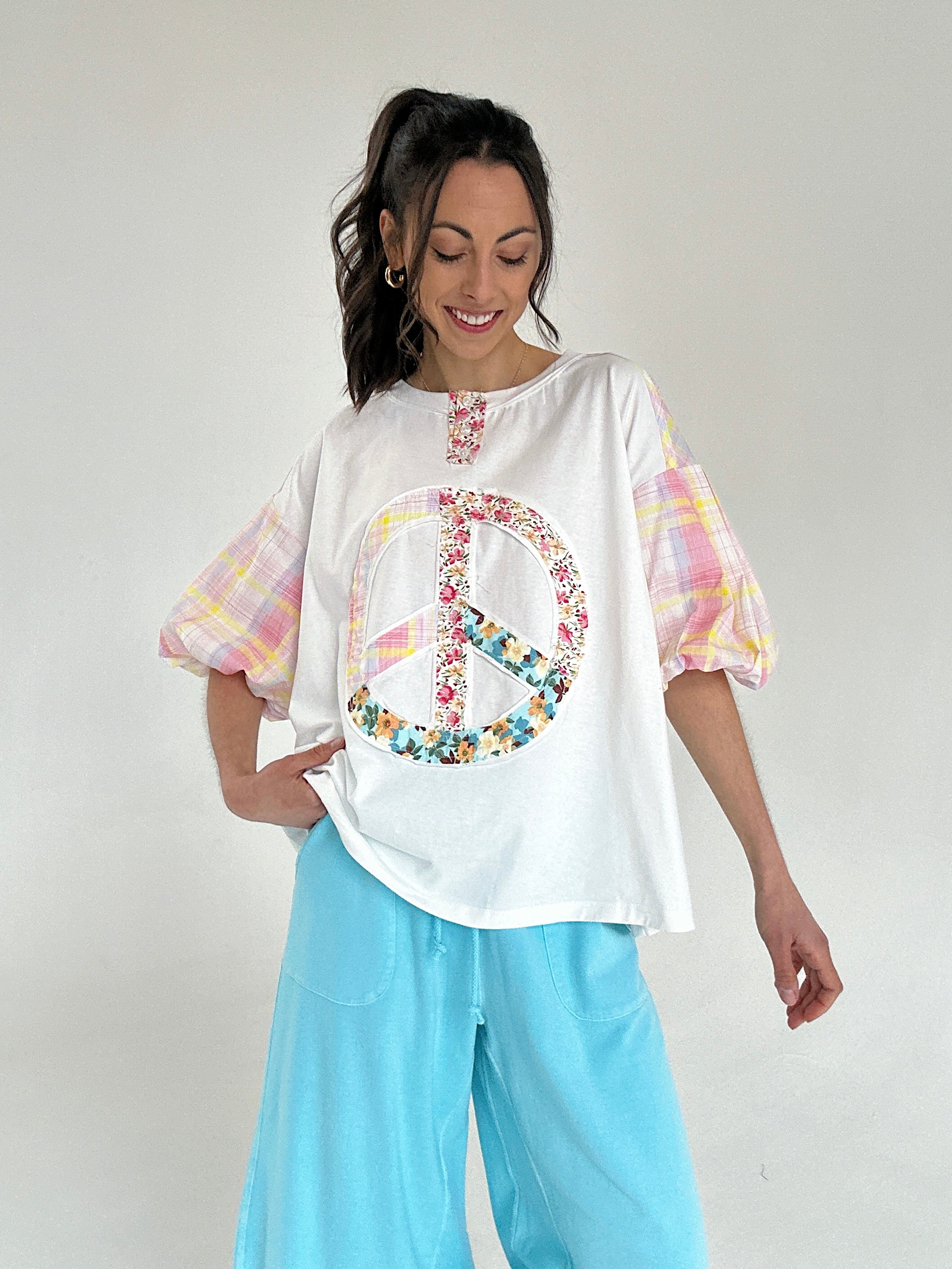 Woman wearing a white tee with a peace symbol design and colorful plaid short sleeves, paired with light blue pants on a plain background.