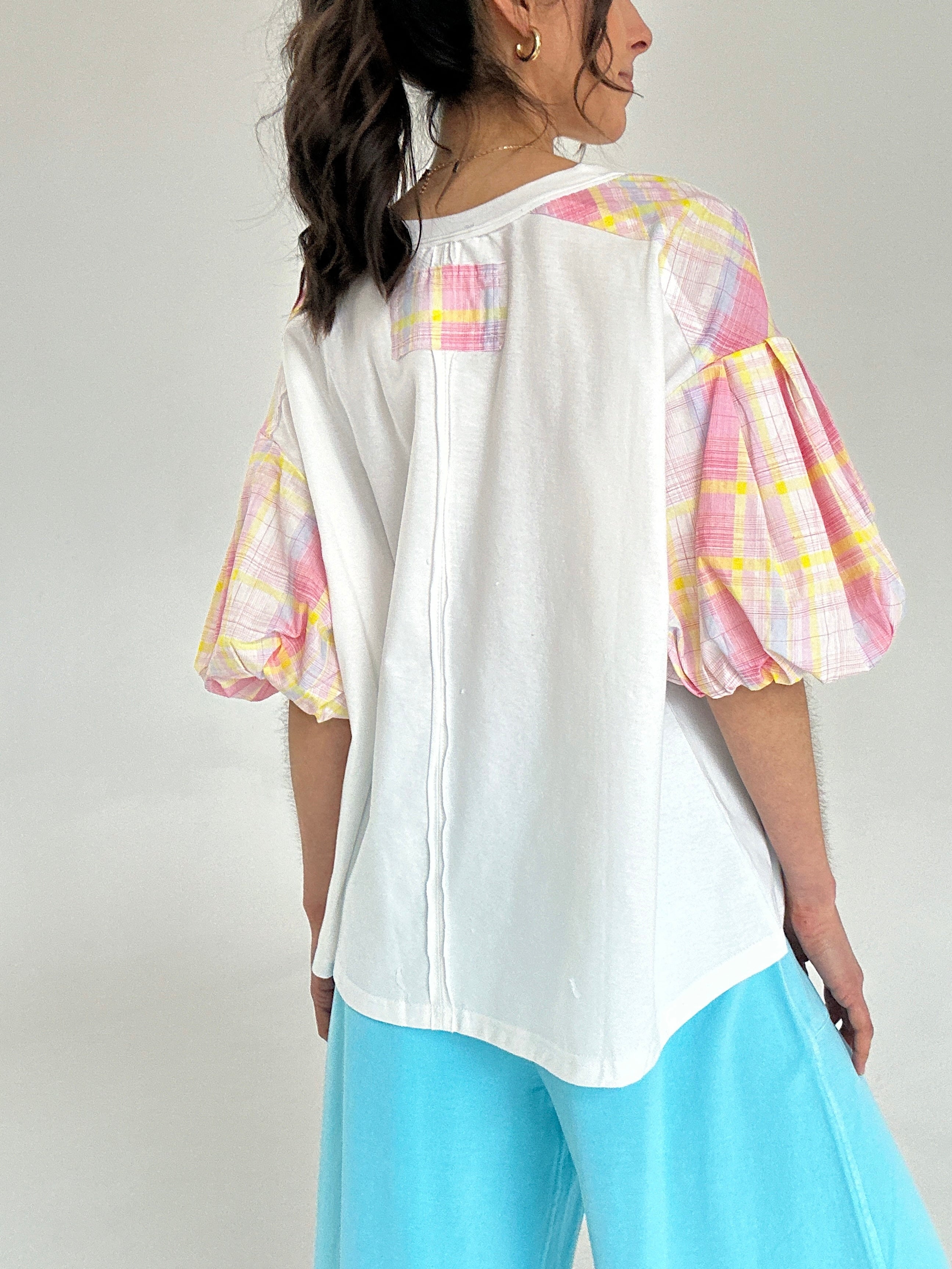 Woman wearing a white blouse with colorful sleeves and light blue pants on a plain background from back
