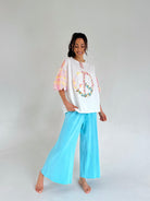 Woman wearing a white top with colorful peace designs and light blue pants on a plain background