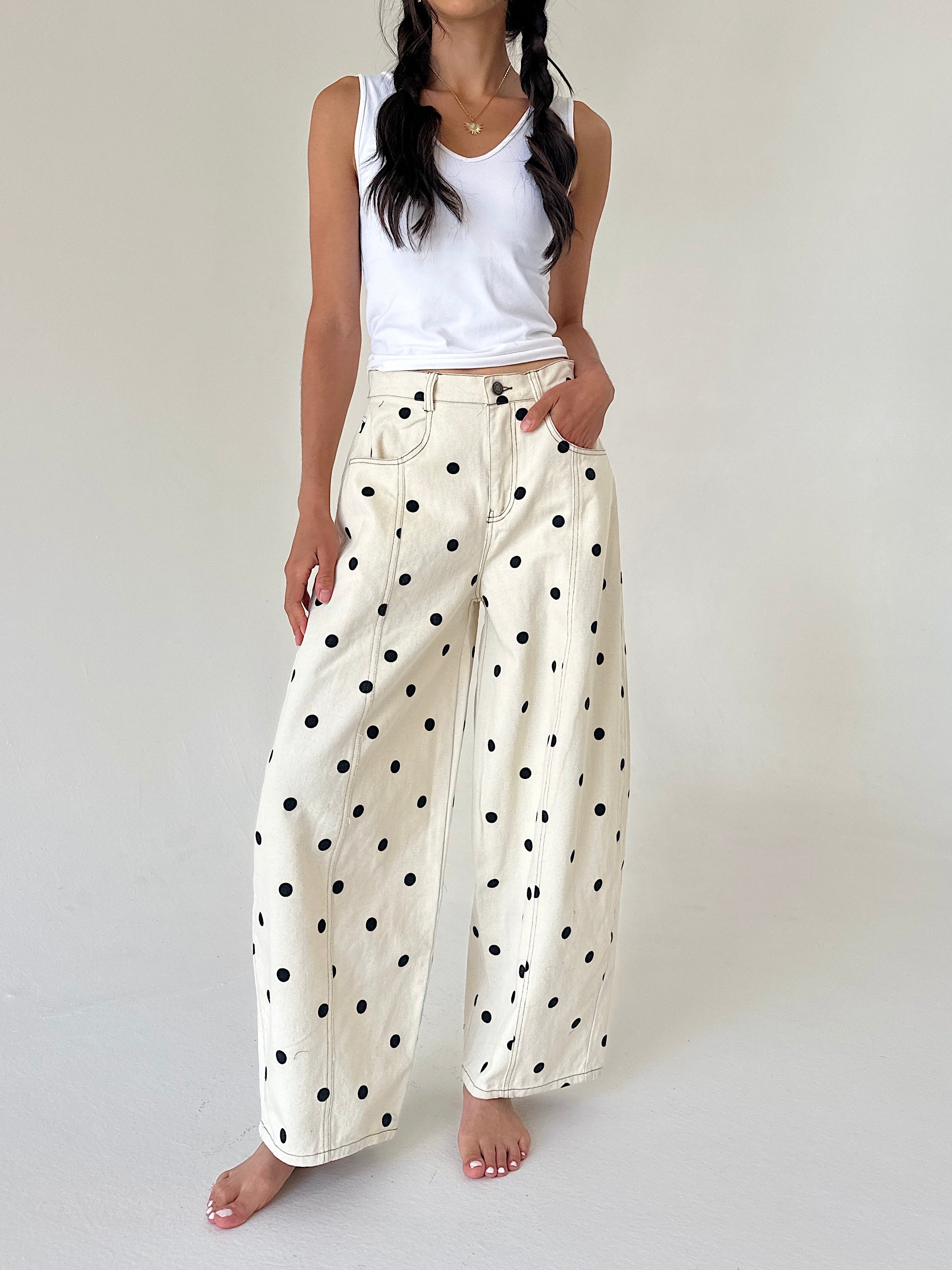 A model wearing cream wide-leg pants with a black polka dot pattern, styled with a white ribbed tank top, standing against a neutral off-white studio background.