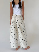 A person shown from the back wearing wide-leg cream pants with black polka dots and a white tank top, standing barefoot against a plain light background.