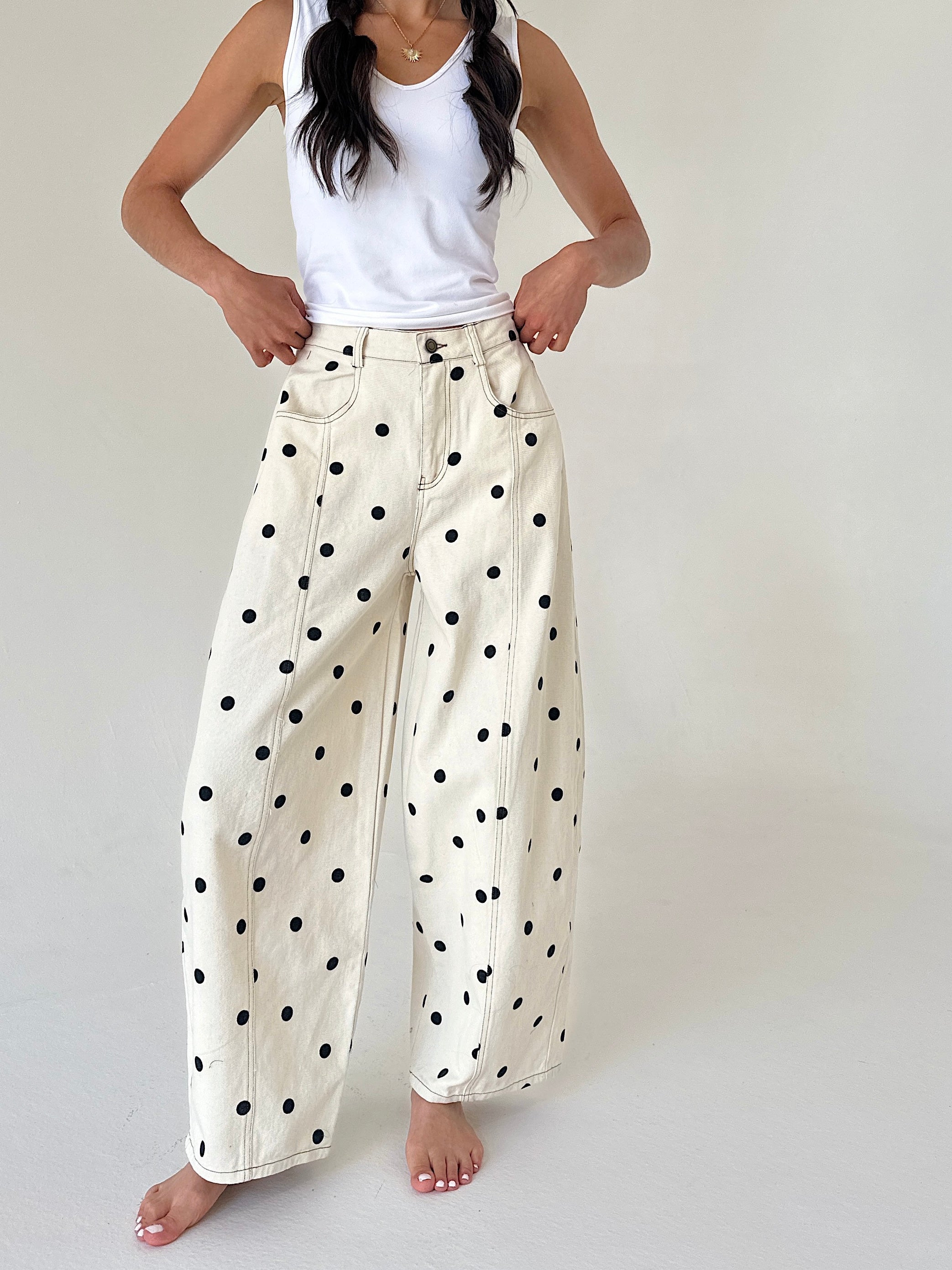 A woman modeling a pair of off-white wide-leg jeans with black polka dots, paired with a white tank top, against a plain light background.