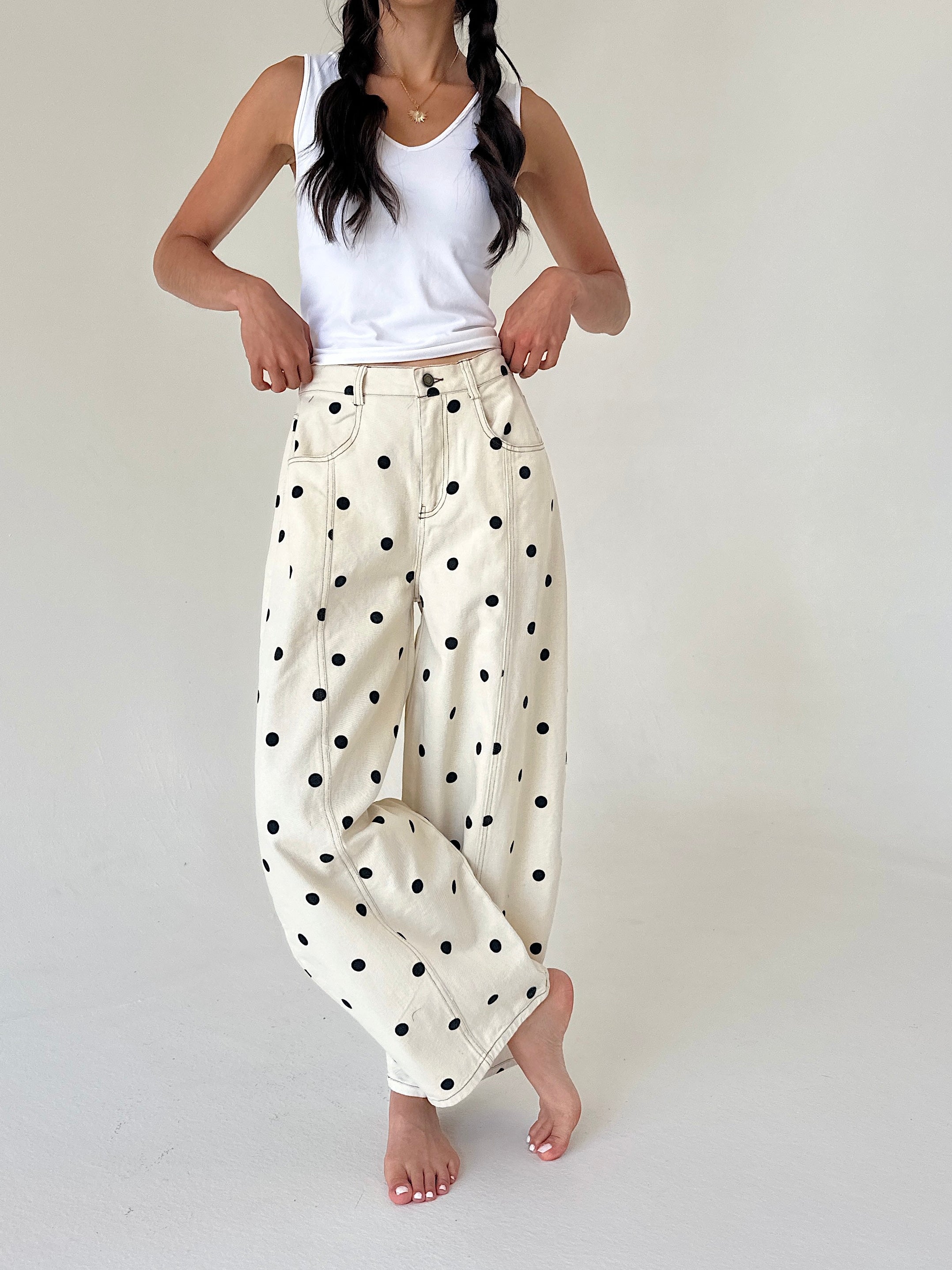 A woman modeling a pair of cream-colored, wide-leg pants with black polka dots, paired with a white tank top, against a plain light background.