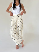 A woman modeling a pair of cream-colored, wide-leg pants with black polka dots, paired with a white tank top, against a plain light background.
