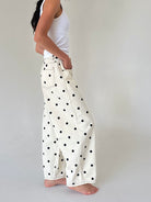 A person wearing off-white wide-leg pants with black polka dots, paired with a white tank top, standing barefoot against a plain light gray studio background.