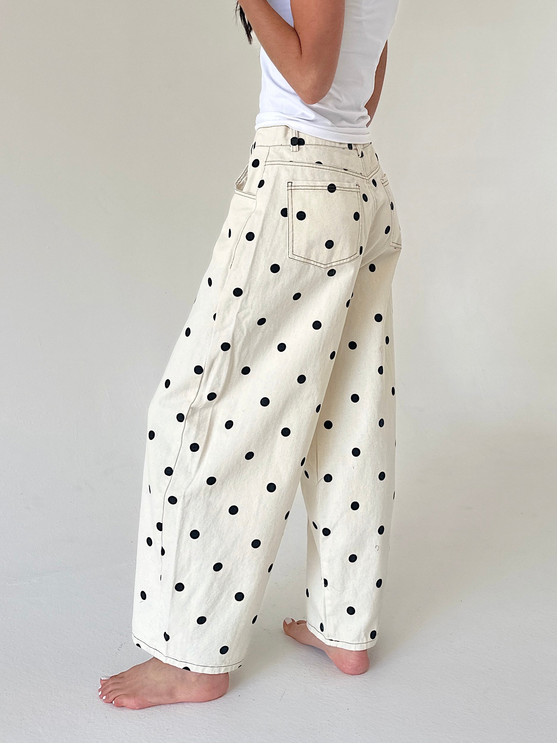 A person shown from the back wearing loose, wide-leg pants in an off-white or cream fabric with a black polka dot pattern, paired with a white tank top, against a plain light gray studio background.