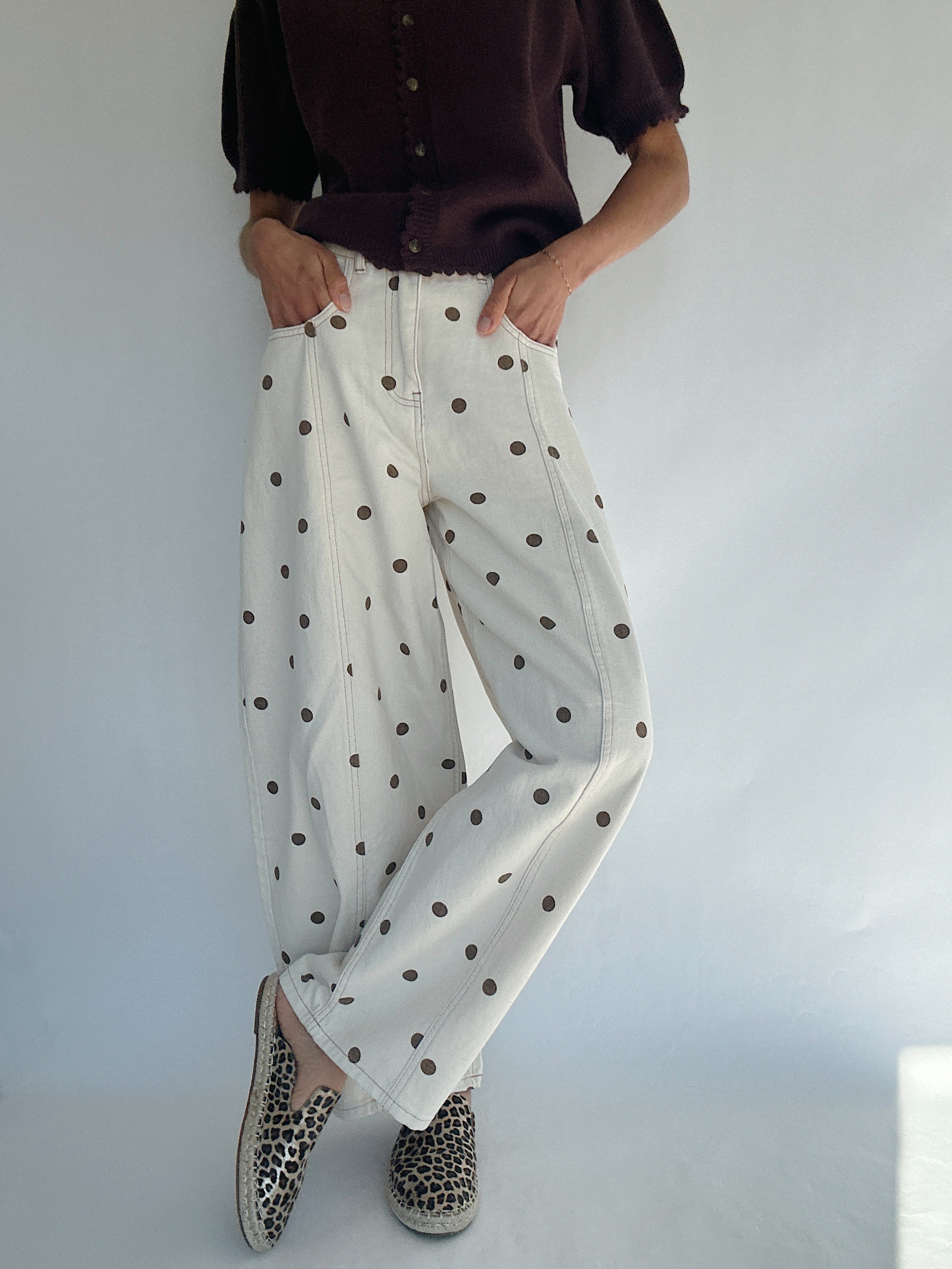Person wearing white barrel style pants with brown polka dots and a brown knit top against a plain background with legs crossed