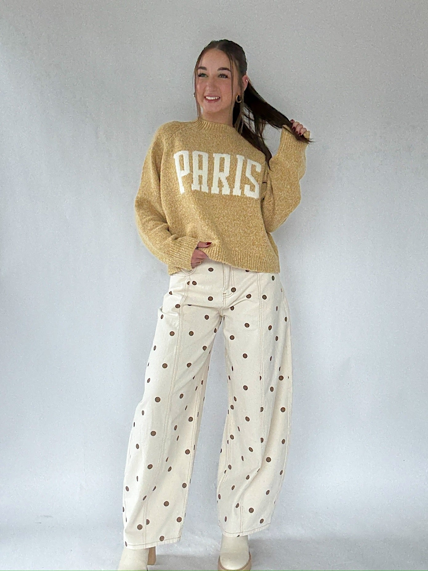 Person wearing a mustard yellow sweater with 'PARIS' text and white polka dot pants on a plain background