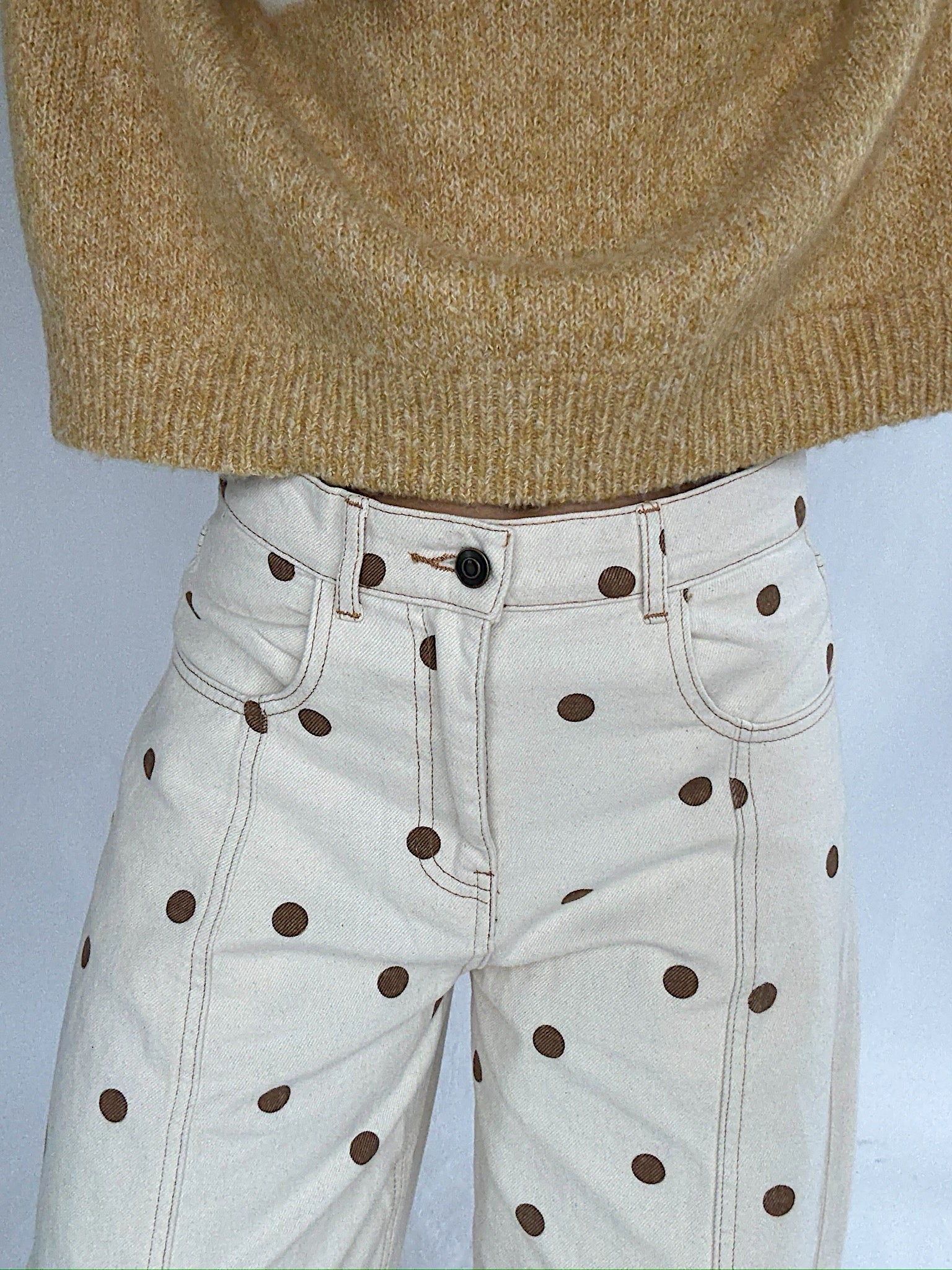 White jeans with brown polka dots worn with a mustard sweater on a light gray background showing off zip and button closure on waistband